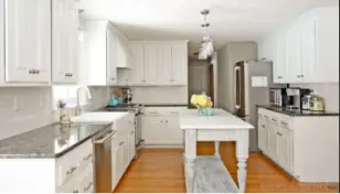 Bright white kitchen with a central island and seating bench, stainless steel fridge, range, microwave, and dishwasher, white cabinets, and pendant lights.