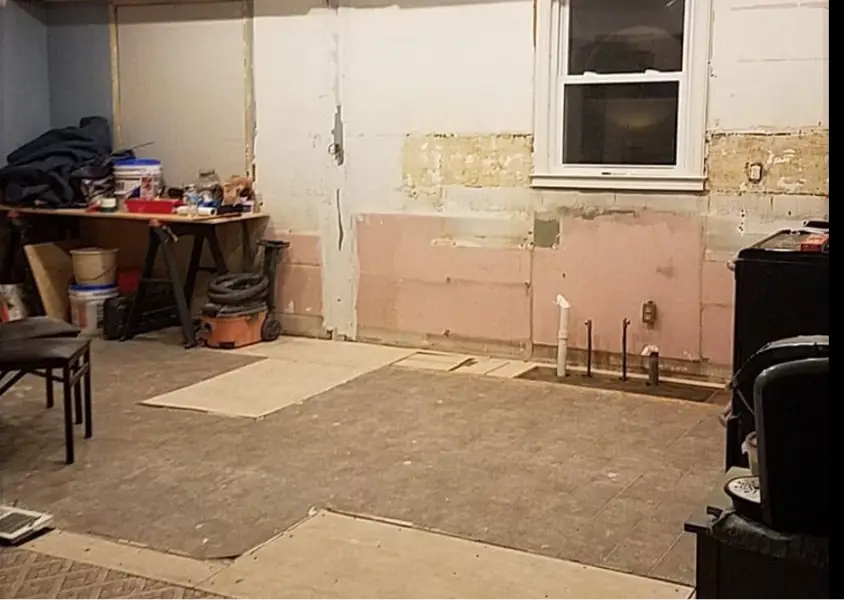 Basement renovation scene with a workbench, tools, and supplies against a wall; exposed pink insulation and a small window.